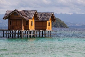 A wooden overwater bungalow with a thatched roof, two bedrooms, and a small porch. It is situated on stilts over clear blue water, with a mountainous backdrop. - Amaris Gorontalo (Gorontalo)