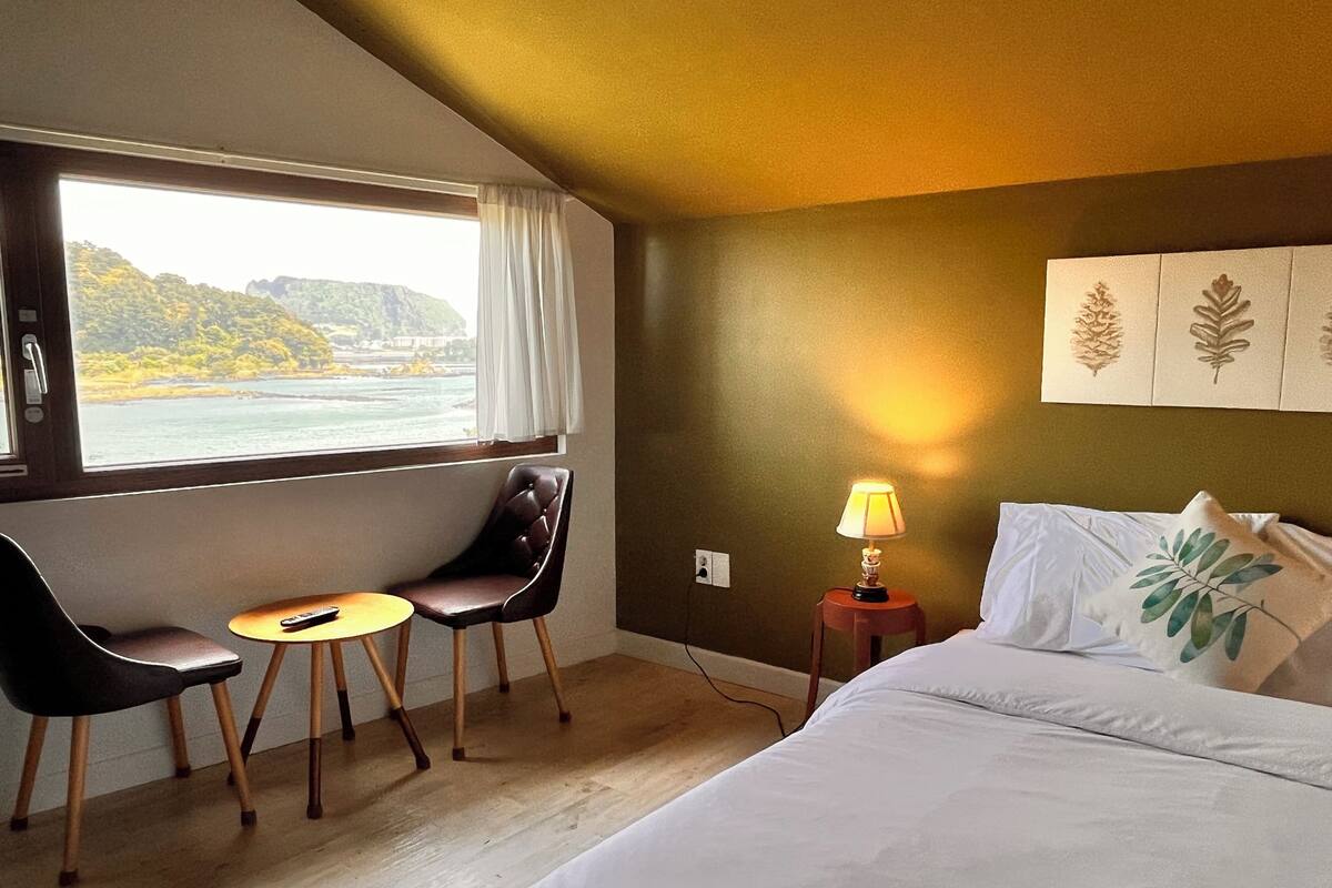 A hotel room with a bed, a desk with a chair, a lamp, and a window with a view of a coastline.