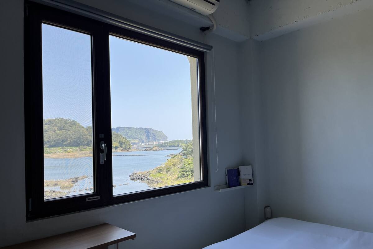 A room with a large window offering a view of a river and hills. There is a bed, a desk, and a shelf with books.