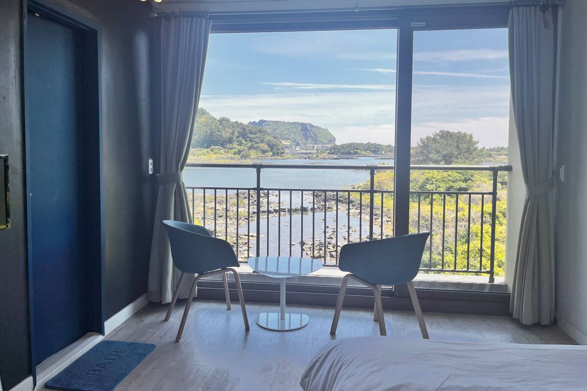 A room with a bed, two chairs, a view of a body of water, and a balcony railing.