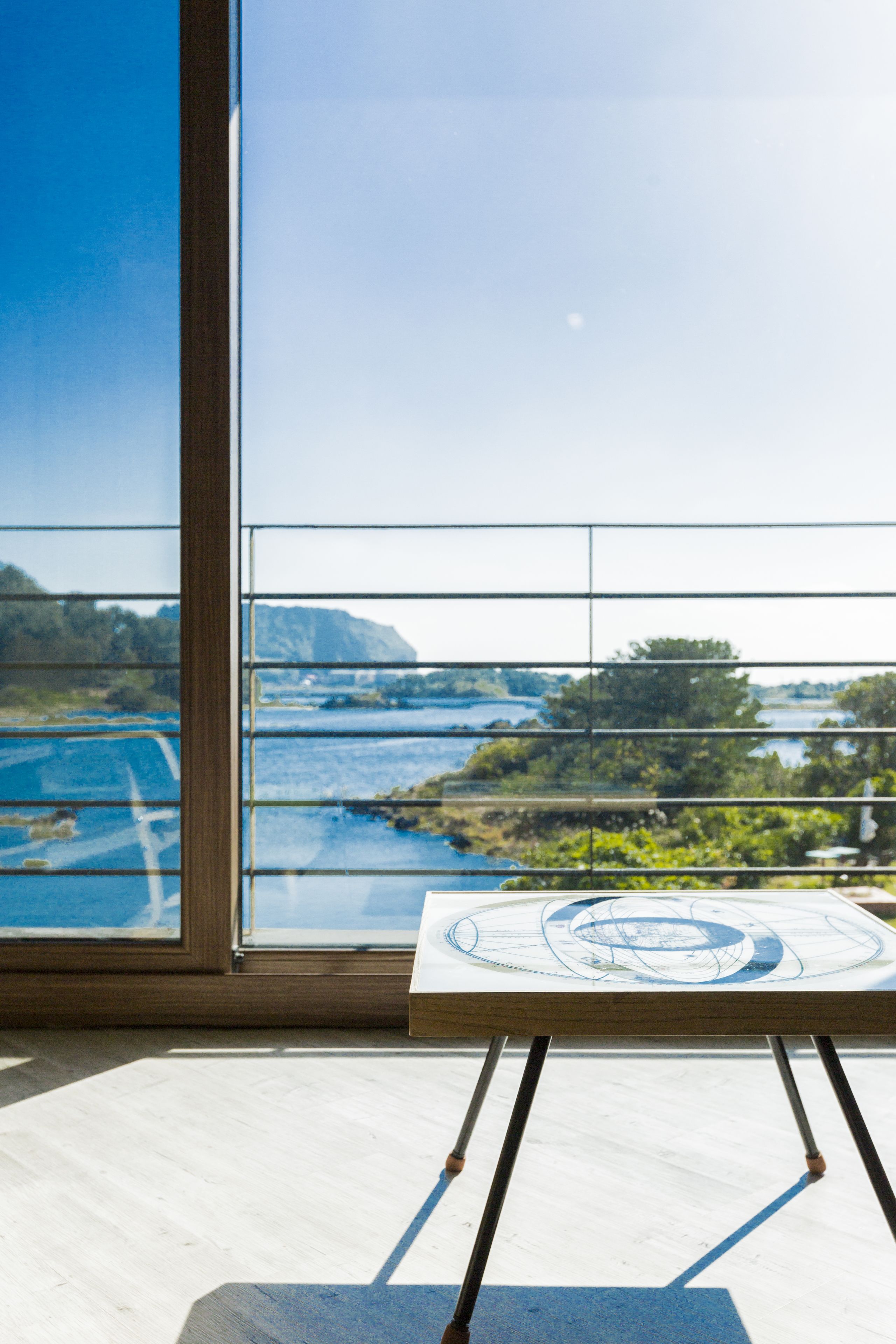 Una habitación con un ventanal grande que ofrece vistas a un paisaje pintoresco, una mesa de madera con un mapa, y una vista de agua y vegetación.