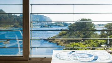 Una habitación con un ventanal grande que ofrece vistas a un paisaje pintoresco, una mesa de madera con un mapa, y una vista de agua y vegetación.