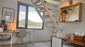 A modern kitchen with a wooden staircase, a large window with a view of greenery, a sink, and a compact refrigerator. - Maison de Ojolac (Seogwipo)