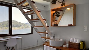 A modern bathroom with a wooden staircase, a large window with a view of a lake, a white sink, and a mirror above it. - Maison de Ojolac (Seogwipo)