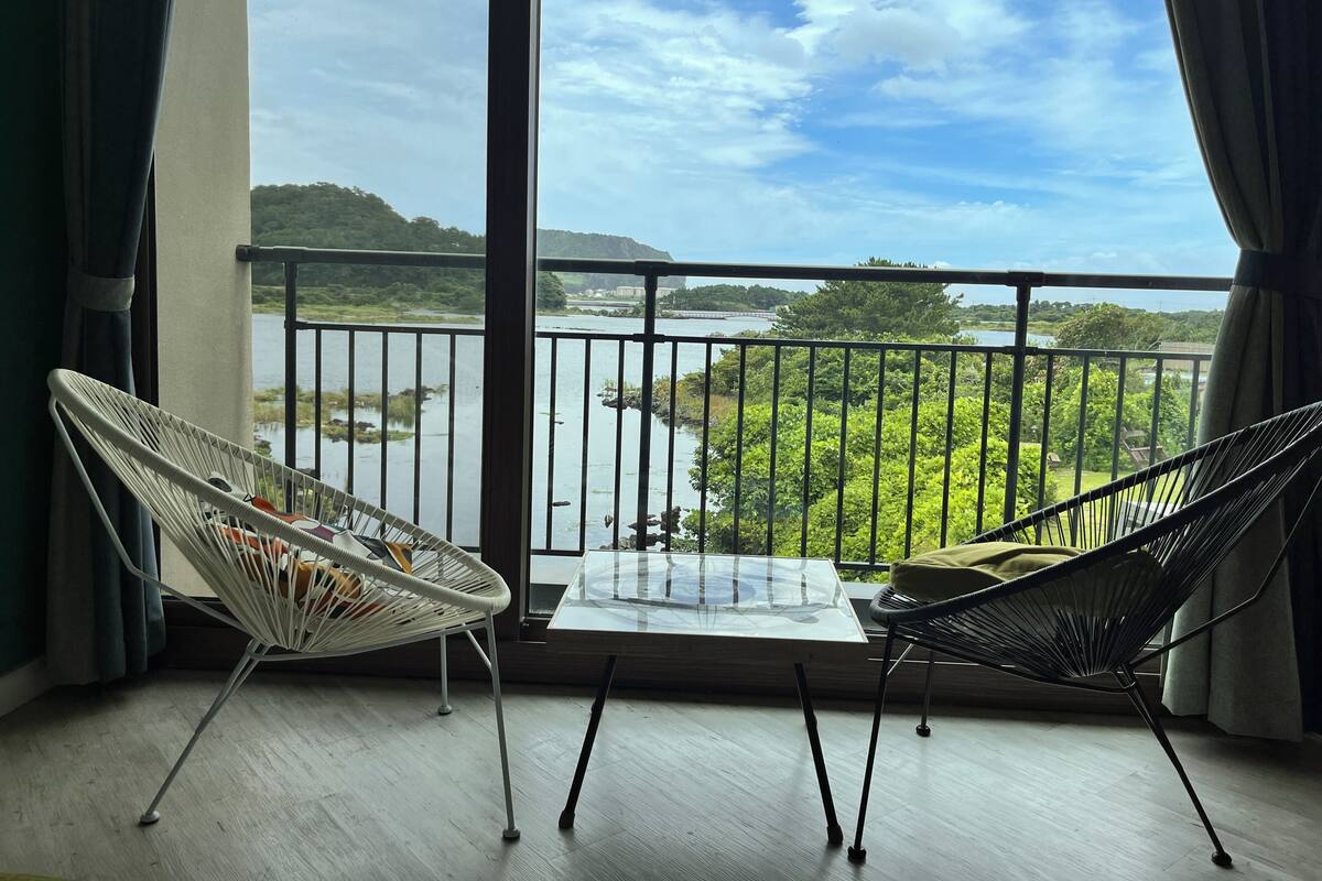 A balcony with two chairs, a glass table, and a view of a river and greenery.