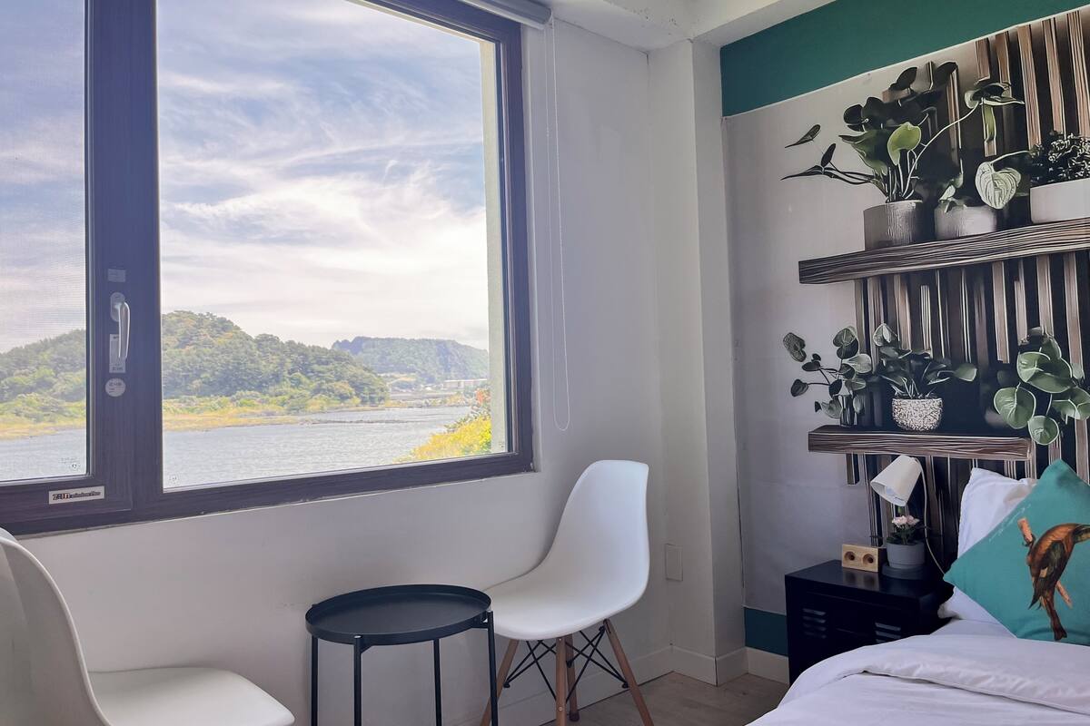A room with a large window, a bed, a white chair, a small table, and a wall-mounted shelf with plants.