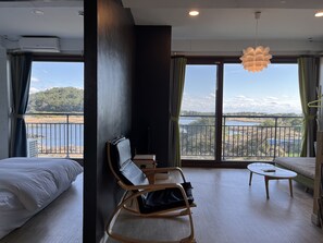 Habitación de hotel con cama, zona de estar con una silla y una mesa, y ventanas amplias con vistas a un lago y un bosque.