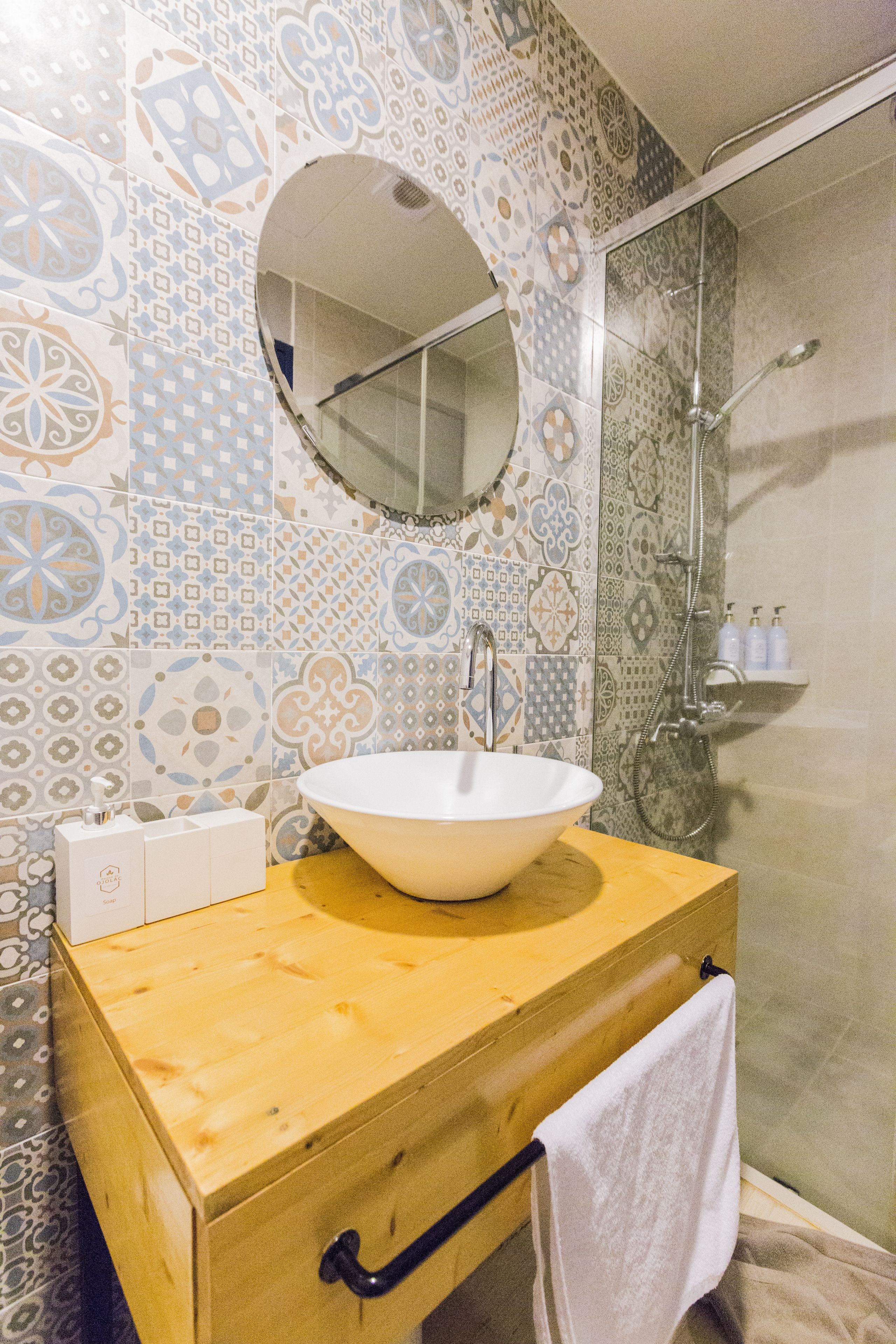 A bathroom with a wooden vanity, a white sink, a round mirror, and patterned tiles.