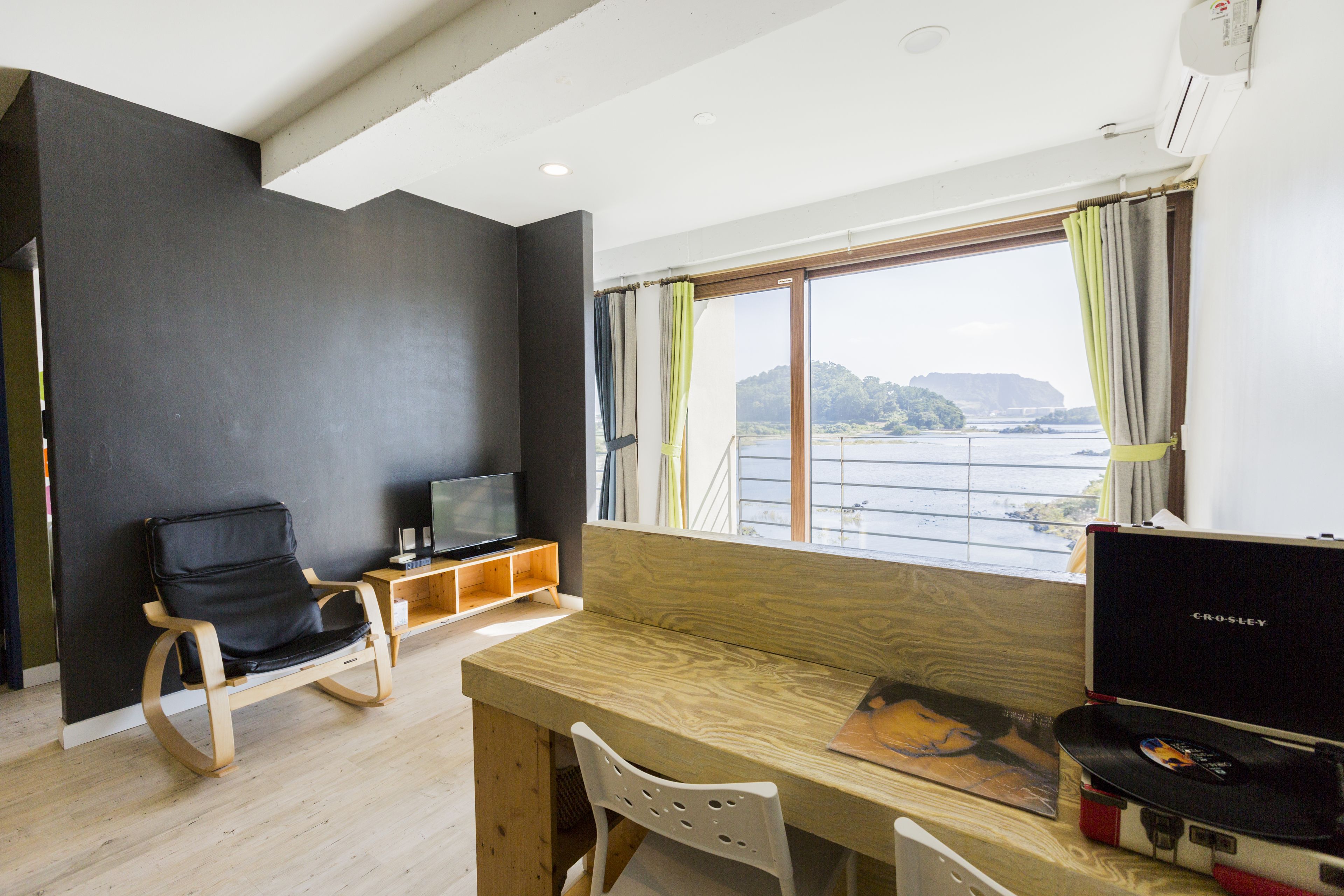 A modern room with a wooden desk, a black leather chair, a television, and a large window with curtains.