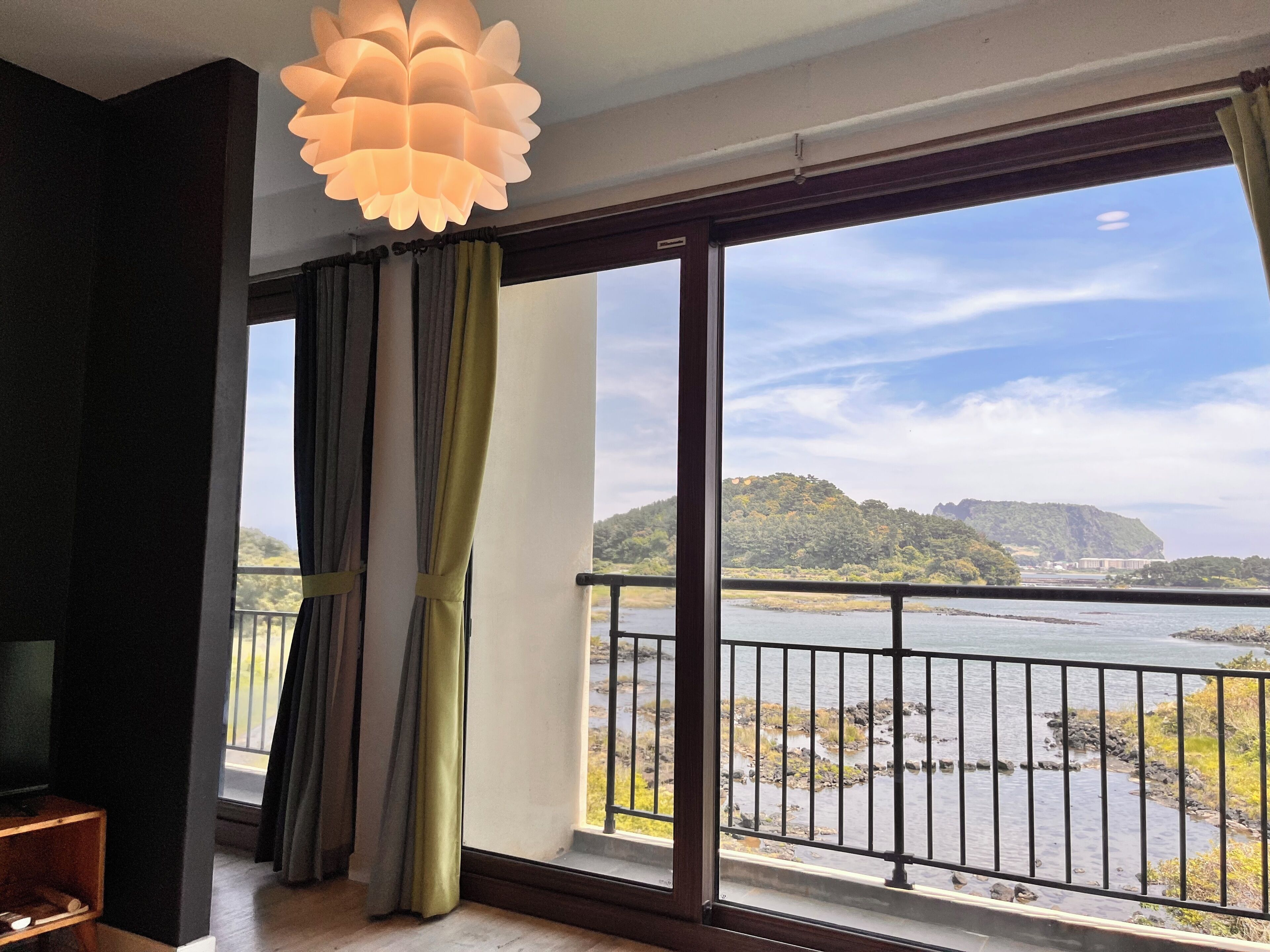 A room with a large window, curtains, a balcony with a railing, and a hanging light fixture.
