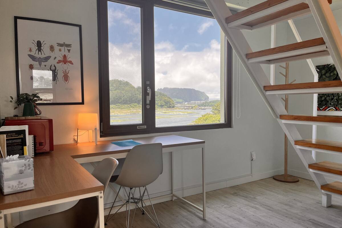 A modern room with a desk, chair, and a large window overlooking a river and mountains.