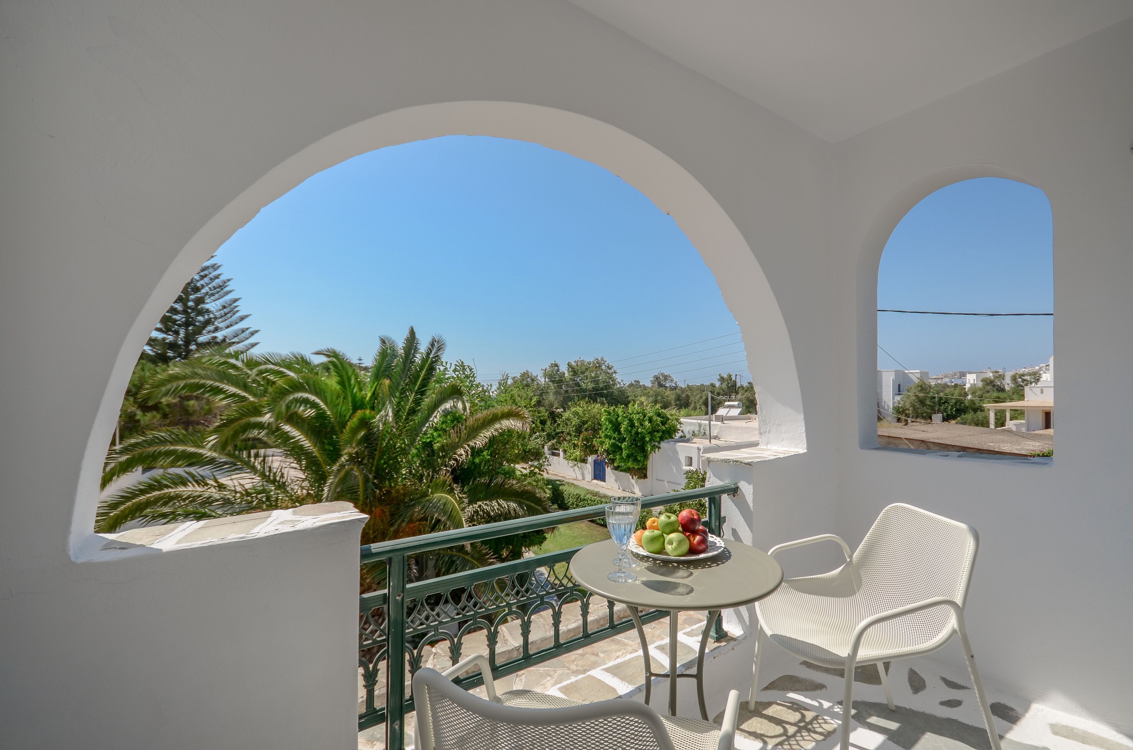 A balcony with white walls and arches, featuring a table with chairs and a fruit bowl.