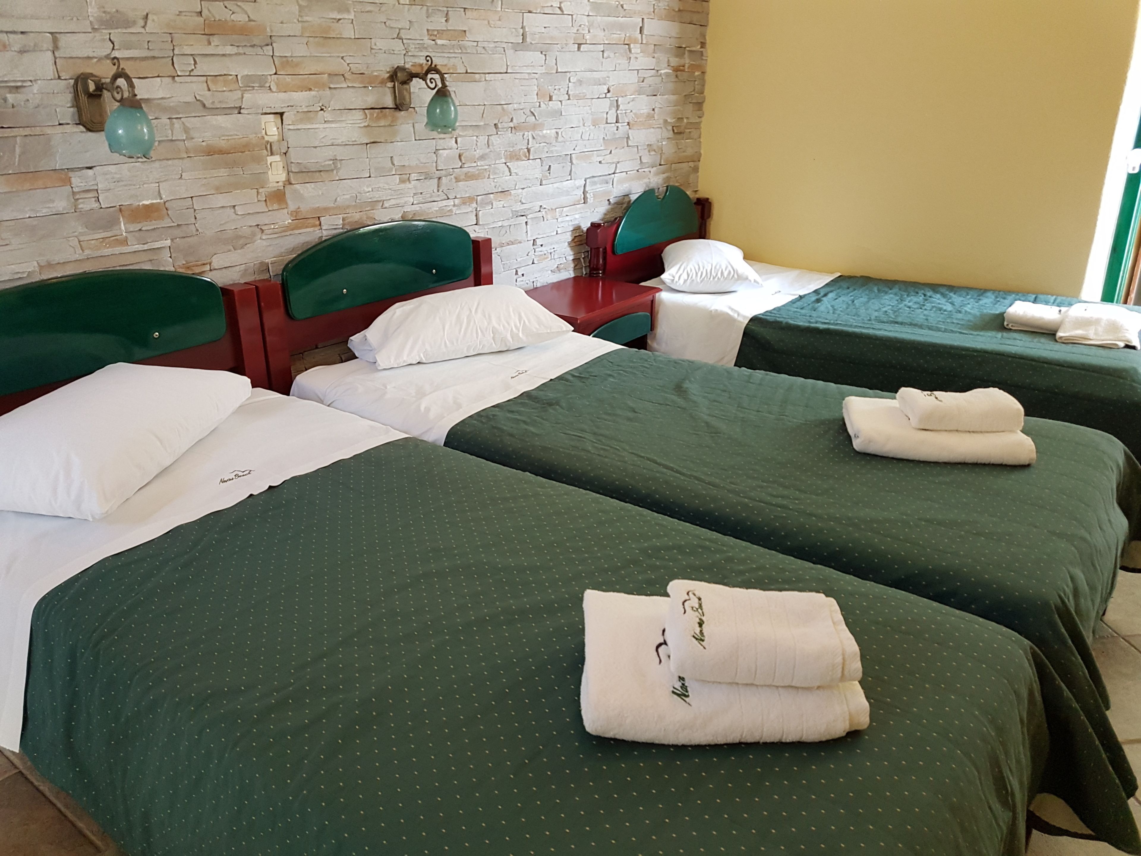 Une chambre d’hôtel avec trois lits, chacun doté de draps verts et d’oreillers blancs. On y trouve deux lampes fixées au mur, avec des abat-jour verts.