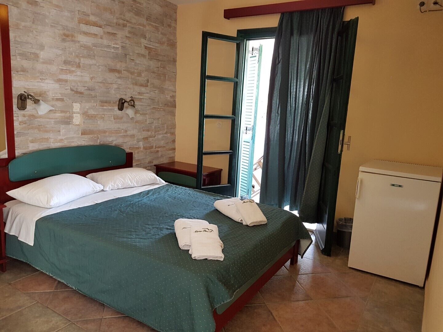 A hotel room with a bed, a small table, a chair, a wardrobe, and a refrigerator.