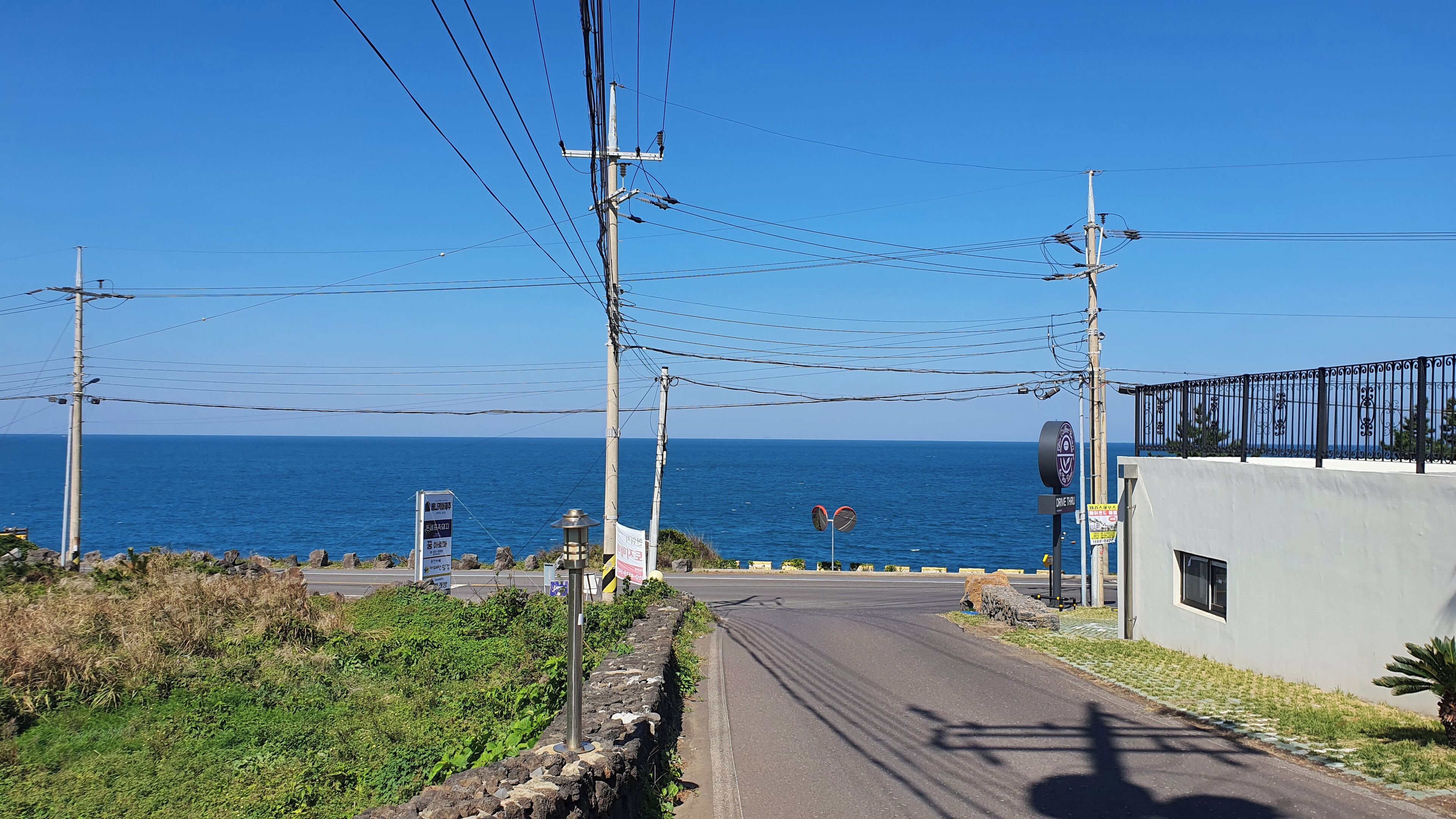 A coastal road with a clear view of the sea, power lines, and a building with a signboard.