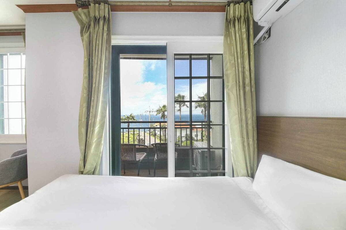 Et hotelværelse med en seng, en stol og en balkon med udsigt over havet og palmer.
