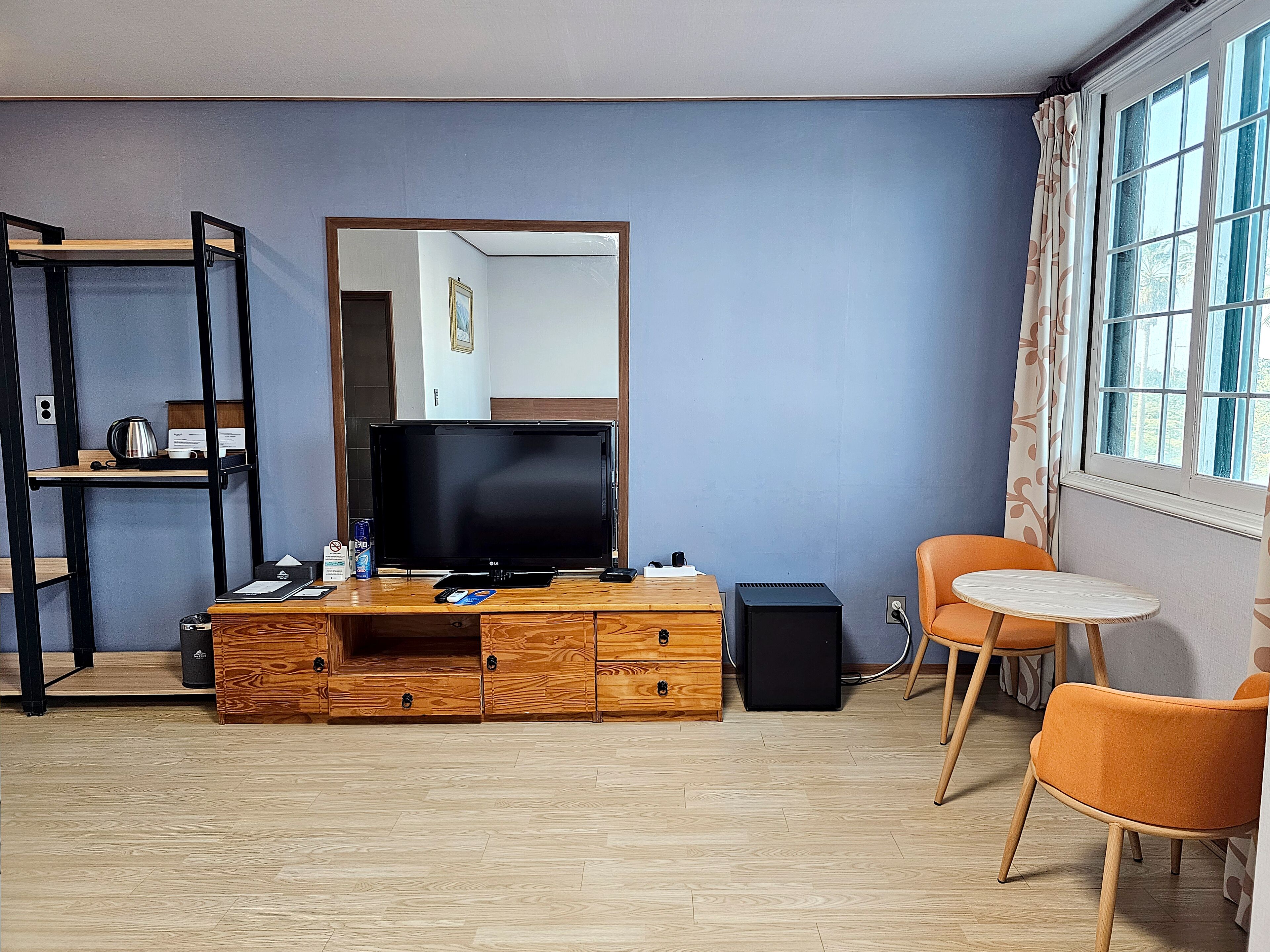 A compact room with a television, wooden storage unit, and a small round table with an orange chair.