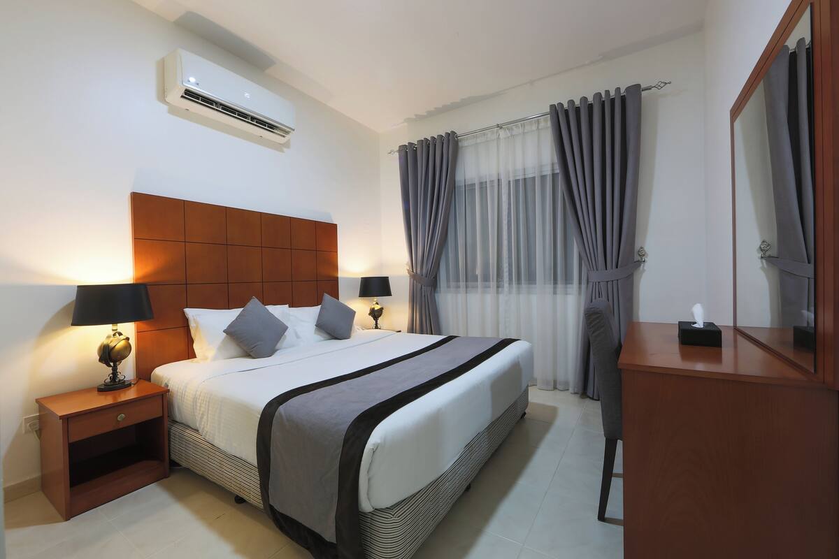 A hotel room with a large bed, two bedside tables with lamps, a wooden headboard, an air conditioning unit, a mirror, and a window with curtains.