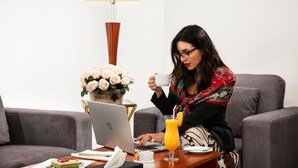 A person sitting on a grey sofa with a laptop, a glass of orange juice, and a white cup on a wooden table. There is a vase with white flowers on the table. - Nova Park Hotel (Sharjah)