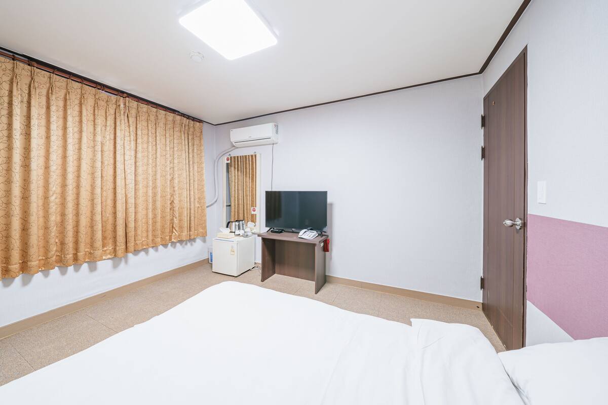A hotel room with a bed, a desk with a computer and phone, a TV, an air conditioning unit, and a door.
