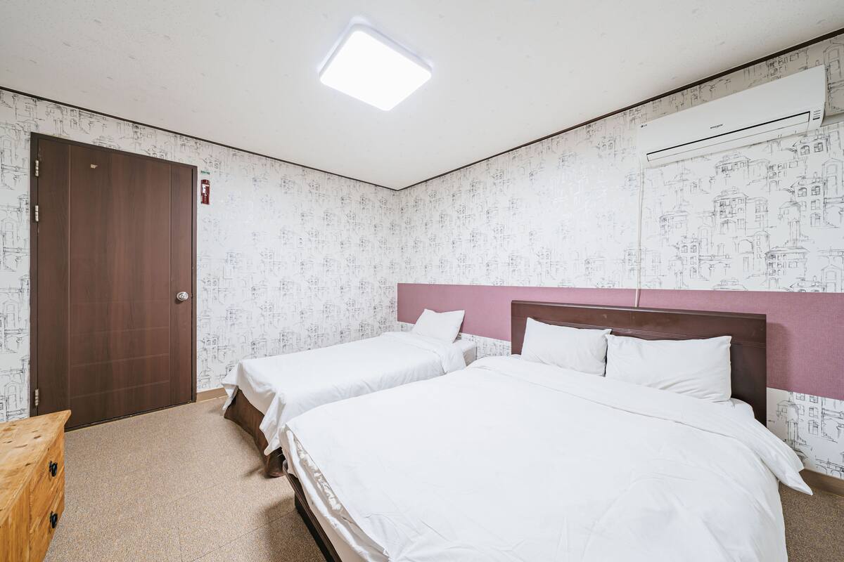 A hotel room with two beds, a wooden door, a ceiling light, and patterned wallpaper.