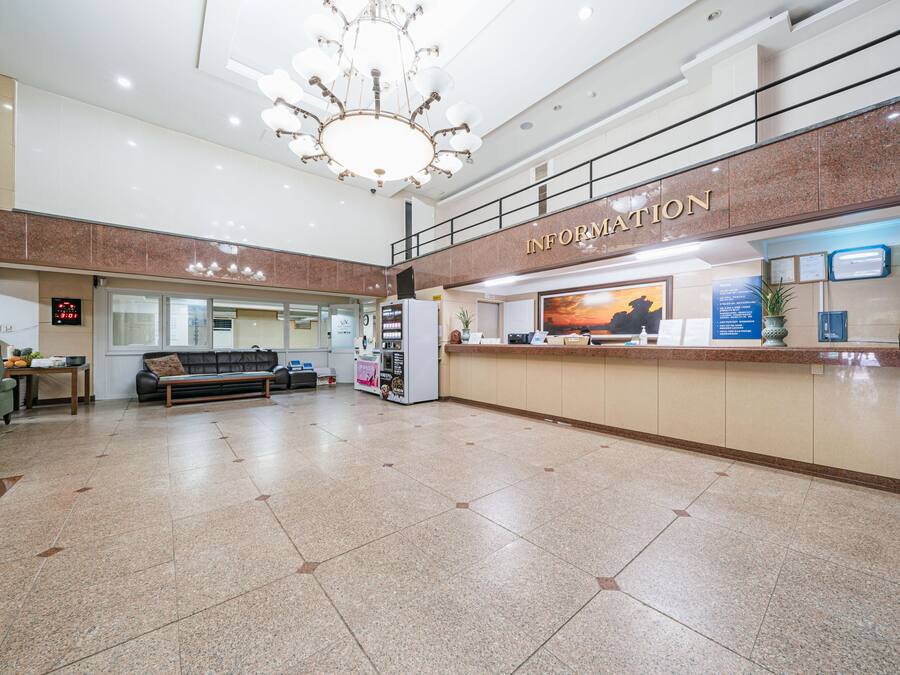 A spacious lobby with a reception desk labeled "INFORMATION," a seating area with a sofa and a coffee machine, and a chandelier.