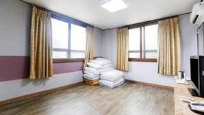 Hotellihuone, jossa on puulattia, kaksi ikkunaa, joissa on verhot, valkoisilla lakanoiden peitetty sänky, työpöytä tietokoneella ja seinälle asennettu ilmastointilaite.
