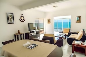 Ein modernes Wohnzimmer mit Essbereich, Fernseher und einem großen Fenster mit Blick auf den Ozean.