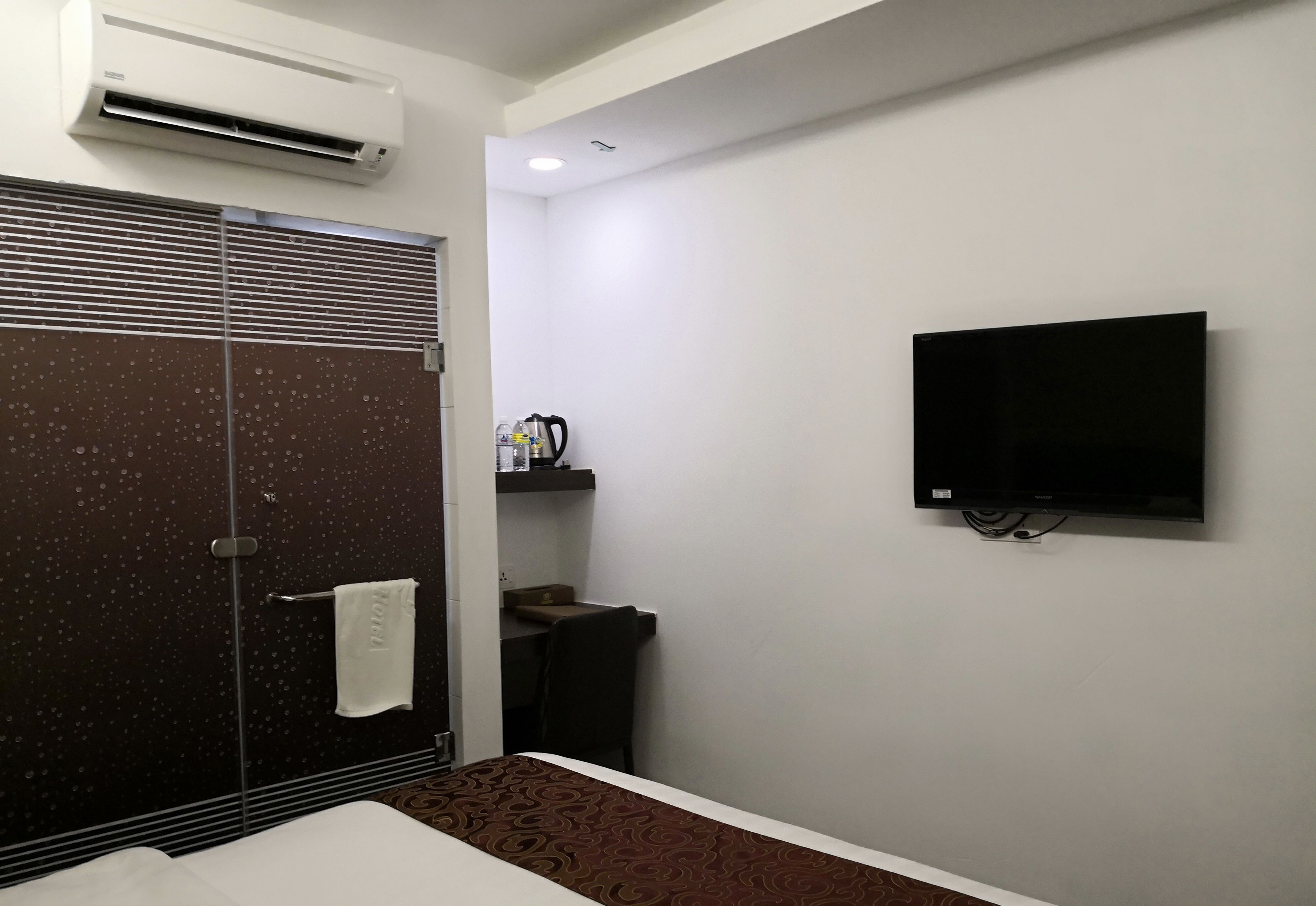 Habitación de hotel con ducha, cama, televisor montado en la pared y una pequeña mesa con tetera y tazas.
