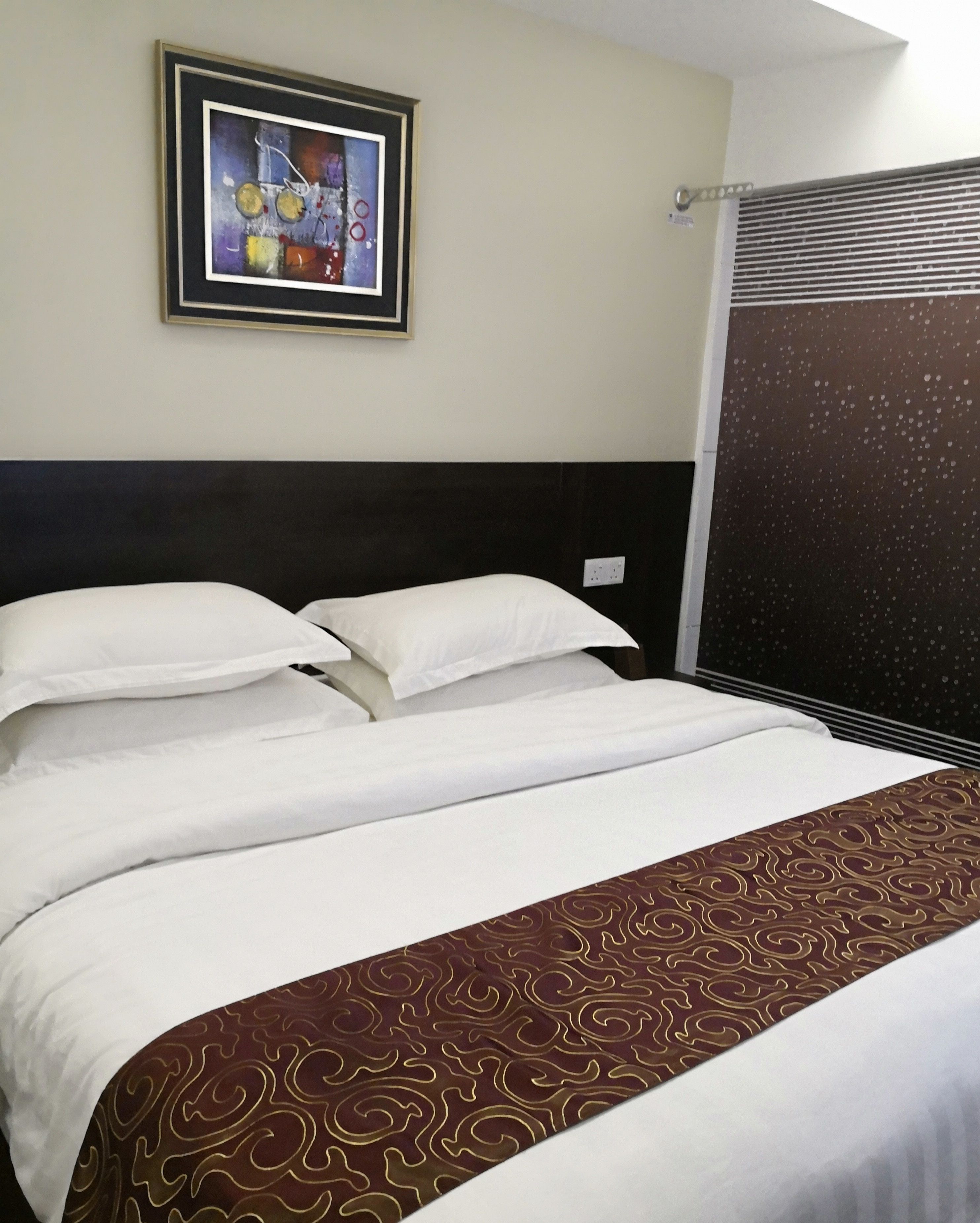 A neatly made bed with white linens and a patterned throw, a framed abstract painting on the wall, and a window with a view of a building.