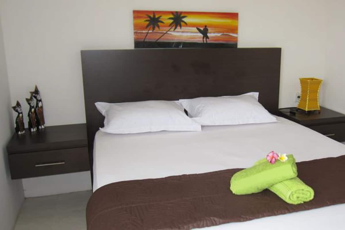 Cama bem arrumada com lençóis brancos e castanhos, um travesseiro decorativo e uma toalha verde com um desenho floral.