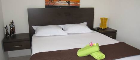 Een net opgemaakt bed met witte en bruine linnens, een decoratief kussen, en een groene handdoek met een bloem.