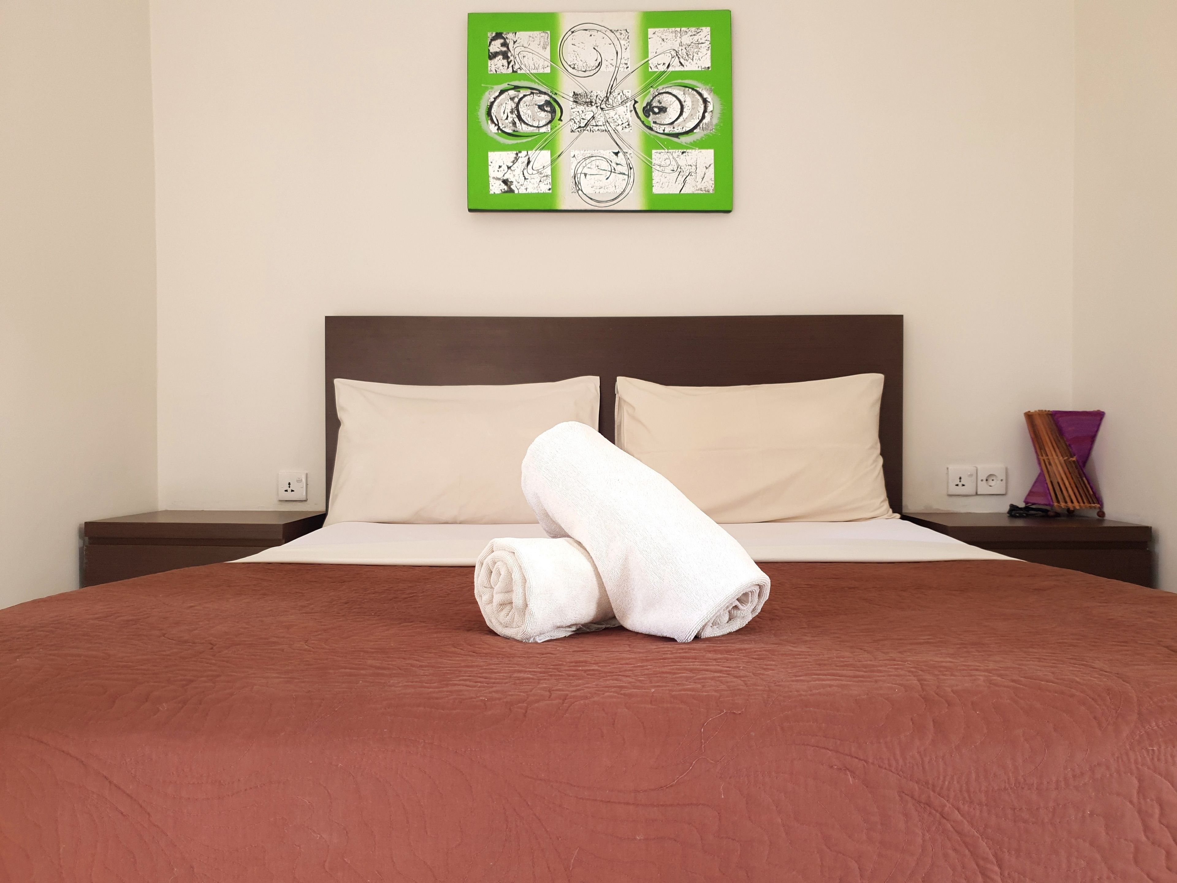 Une chambre d’hôtel avec un grand lit, deux oreillers, une serviette pliée, une œuvre d’art abstraite verte et une table de chevet avec une lampe.