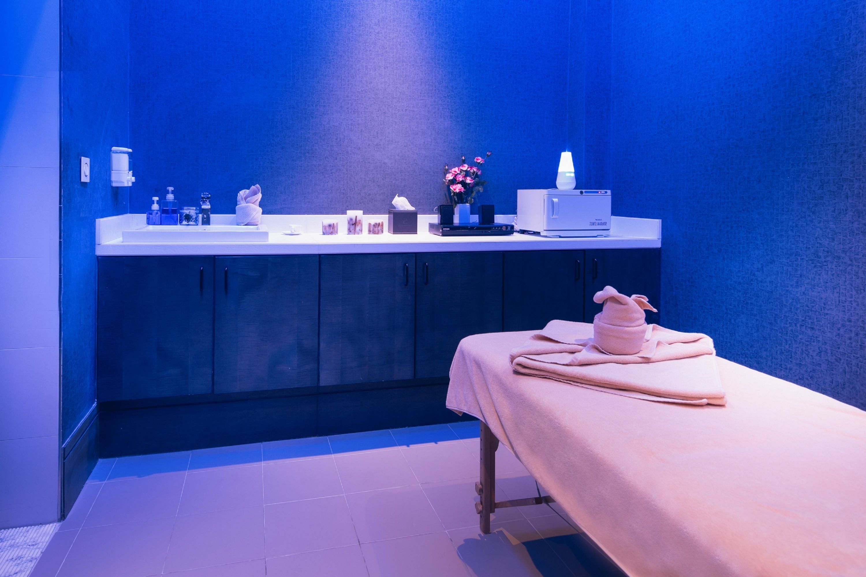 A modern bathroom with a blue wall, a white sink, and a massage table with towels.