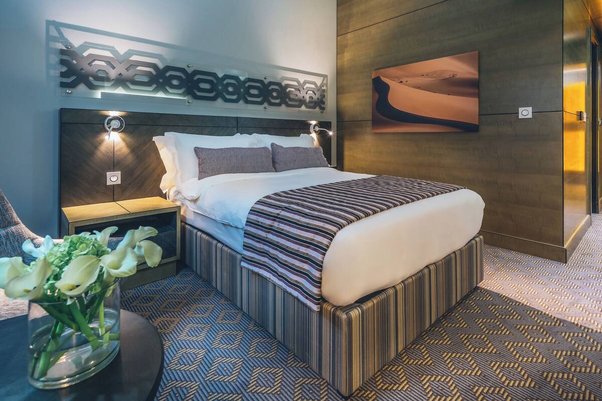 A modern hotel room with a large bed, a bedside table with a vase of flowers, a wall-mounted artwork, and a patterned carpet.