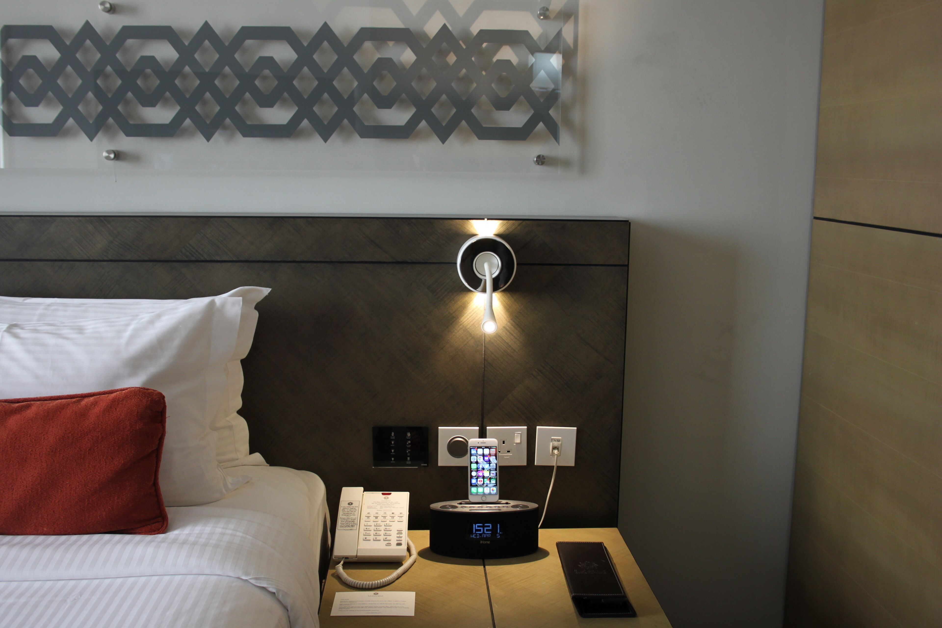 A hotel room with a bed, a bedside table with a phone and a charging station, and a wall-mounted light fixture.