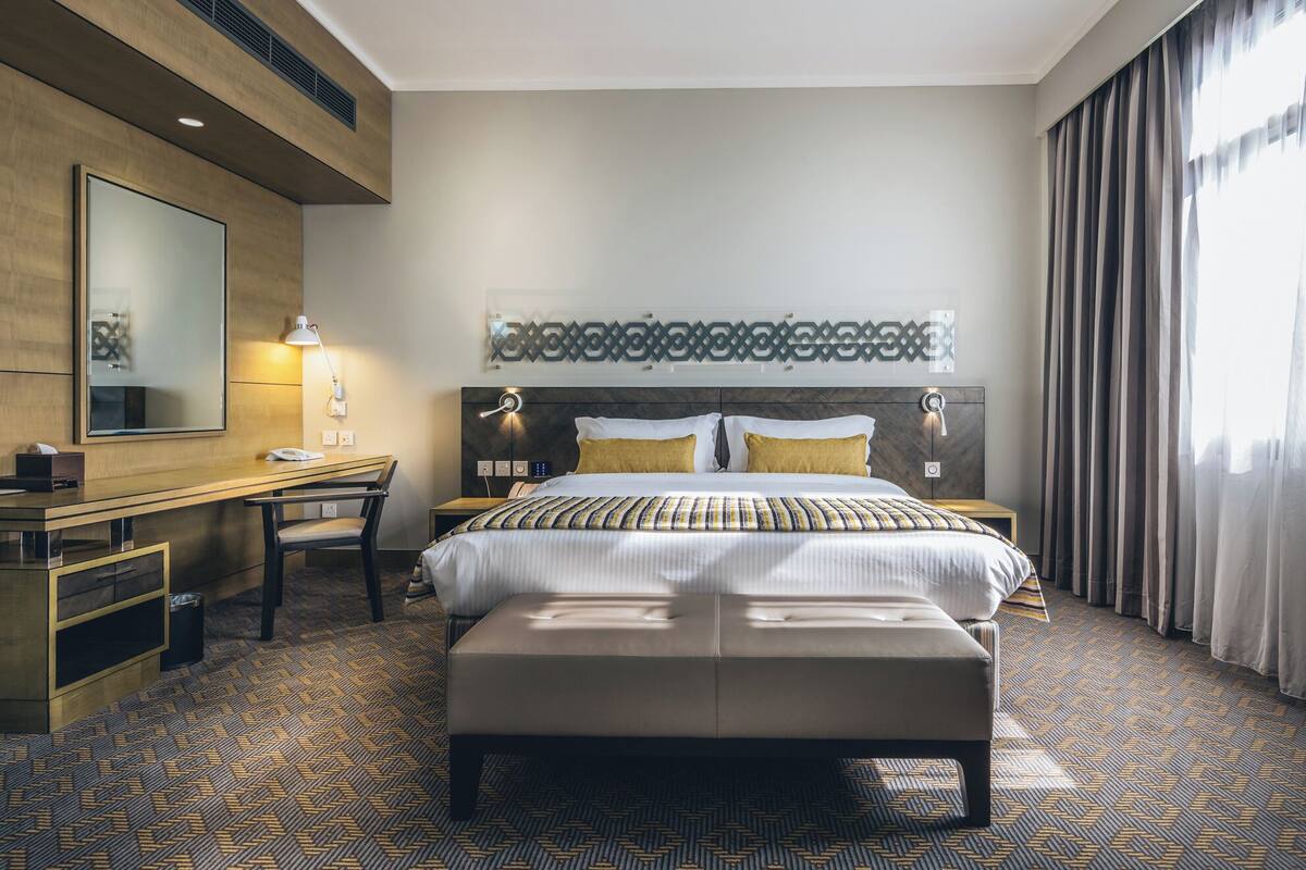 A hotel room with a bed, a desk, a chair, a bench, a mirror, and a window with curtains.