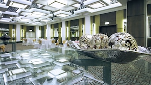 Un moderno vestíbulo de hotel con una mesa de vidrio que exhibe cubiertos ornamentados, un piso con diseño geométrico e iluminación empotrada en el techo.