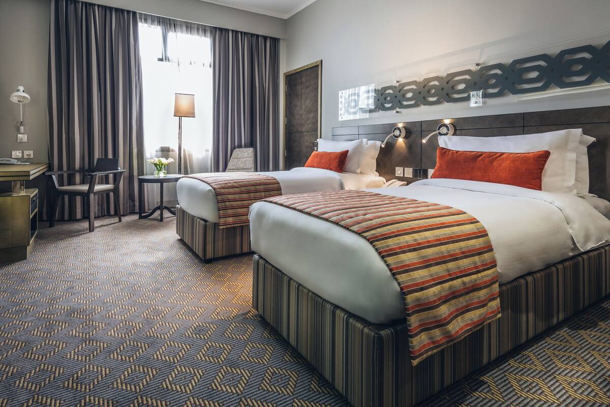 A hotel room with two beds, a desk, a chair, a lamp, and a large window with curtains.