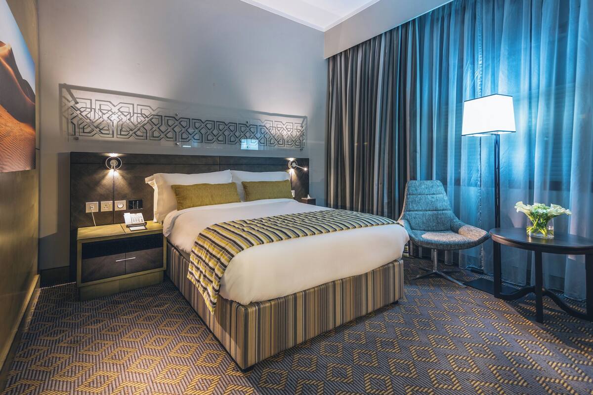 A hotel room with a large bed, a nightstand with a lamp, a chair, a small table with a vase of flowers, and a view of the outside through sheer curtains.