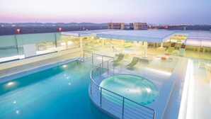 Un área de piscina en la azotea con sillones de descanso y un jacuzzi, rodeada de barandas de vidrio y una vista despejada del paisaje urbano al atardecer.