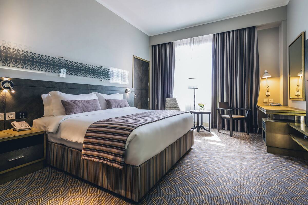 A hotel room with a large bed, a desk, a chair, a small table, and a mirror.