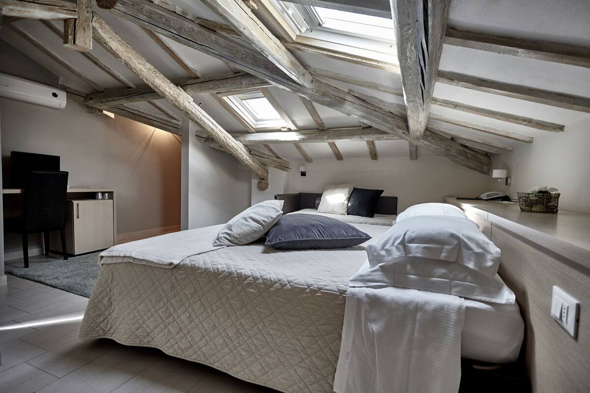 Une chambre avec un plafond en pente, des fenêtres de toit, un lit avec des oreillers, une table de chevet et un bureau avec une chaise.