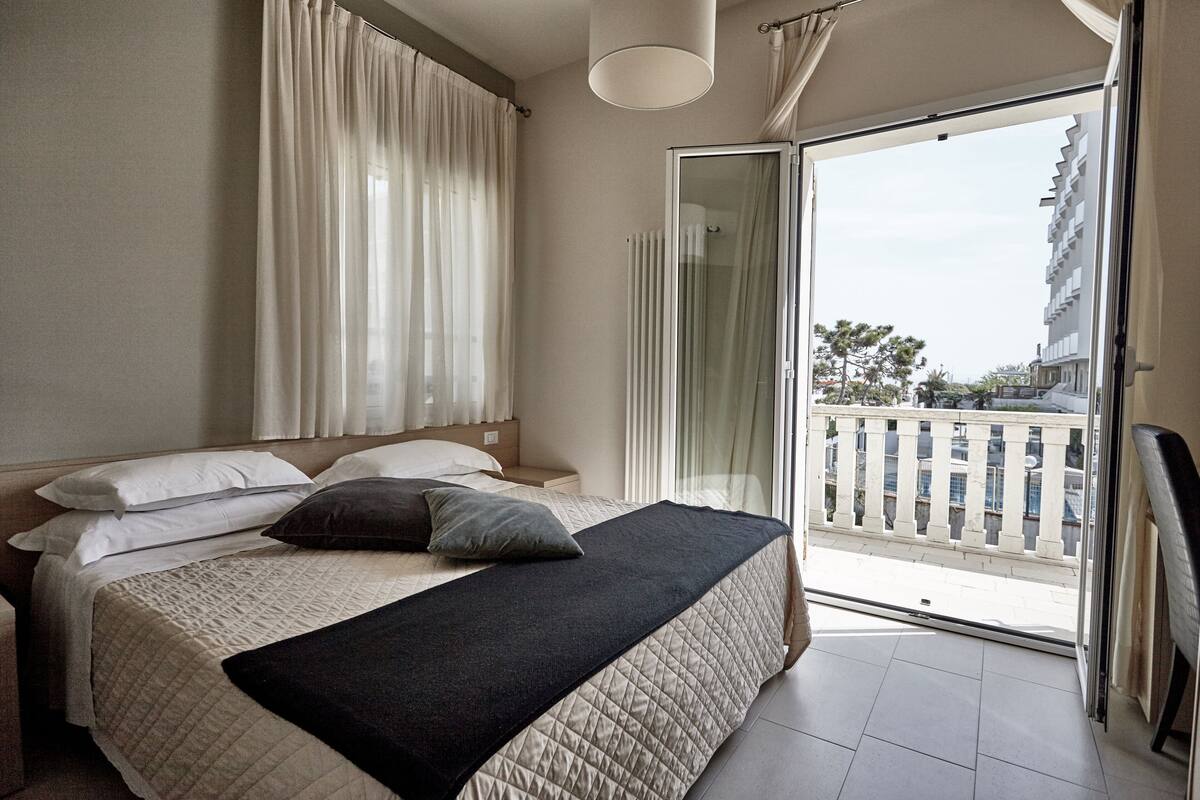 Une chambre à coucher avec un lit, des rideaux, un balcon avec une balustrade et une vue sur les bâtiments.