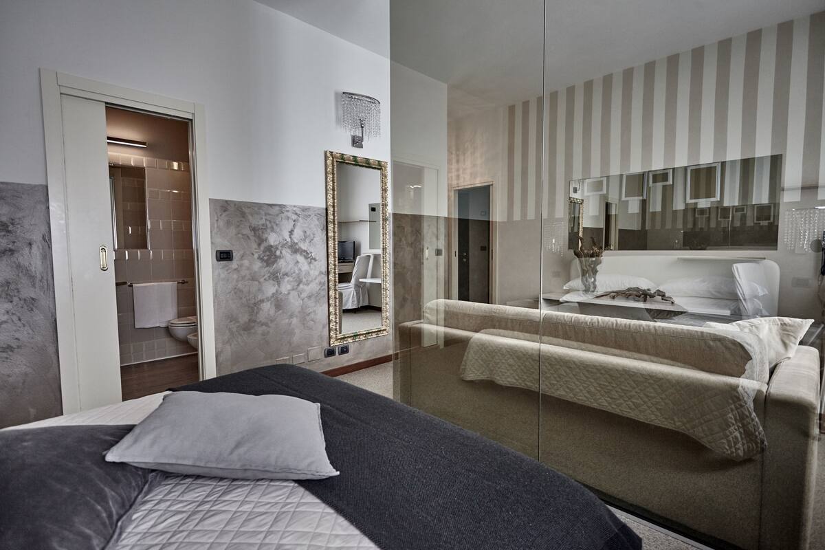 Une chambre d’hôtel moderne dotée d’un grand lit, d’une cloison en verre, d’un mur miroir et d’un coin salon séparé avec une table de massage.