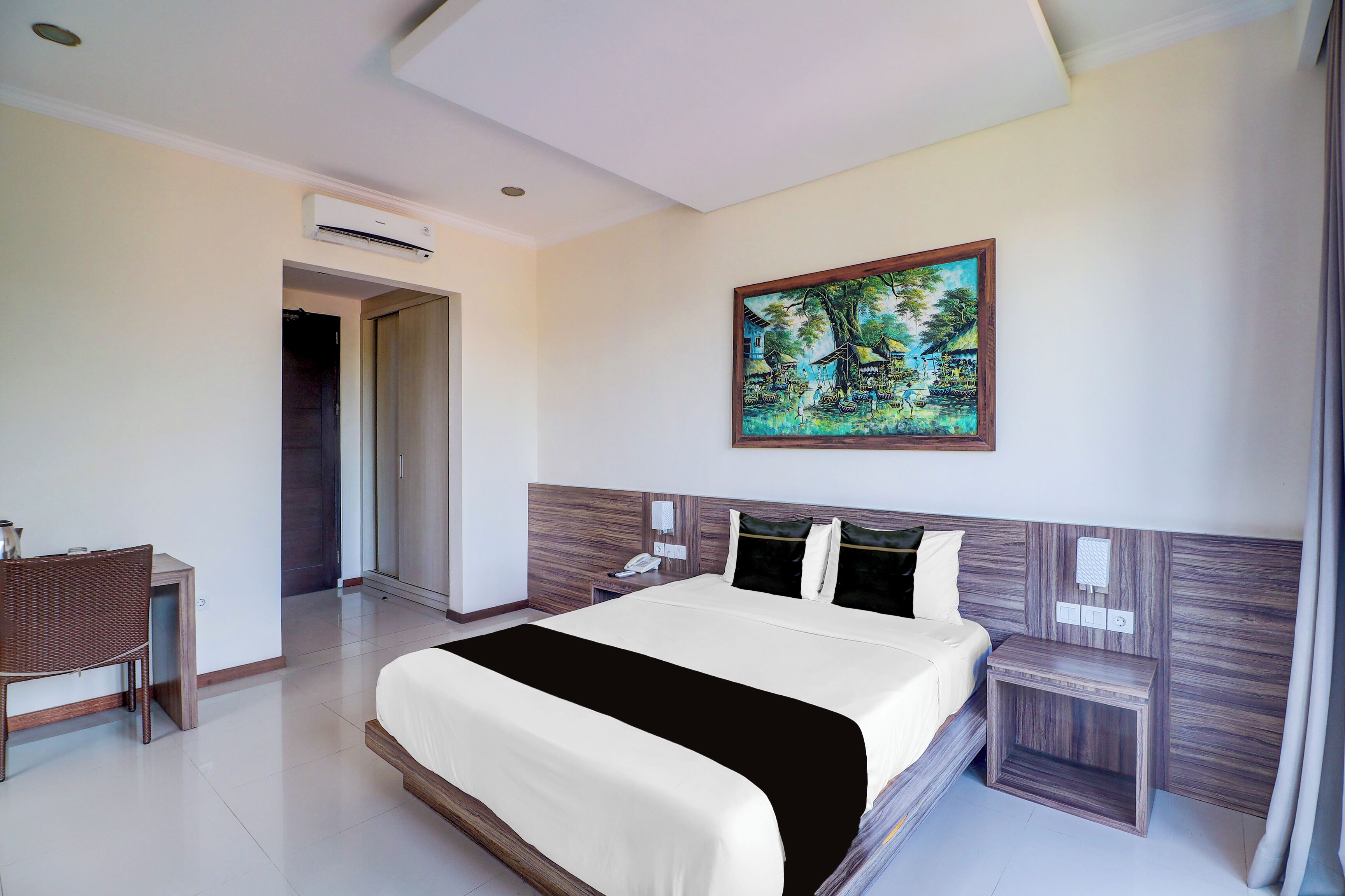 A hotel room with a bed, a desk, a chair, an air conditioning unit, and a painting on the wall.