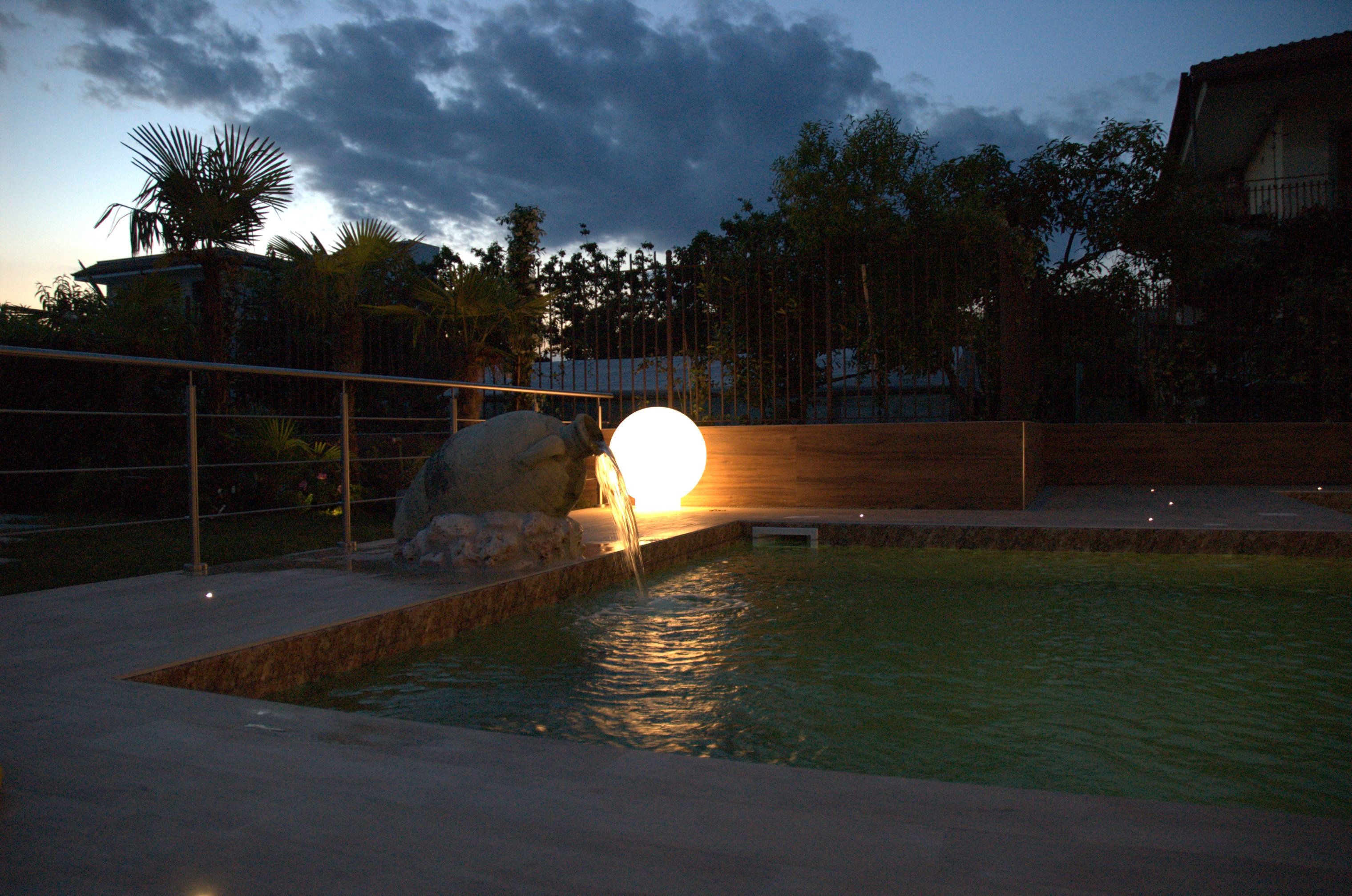 A swimming pool with a waterfall feature, surrounded by a fence and palm trees.