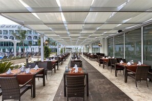 A covered outdoor dining area with tables and chairs, large windows, and a view of a building in the background. - Karmir Resort & Spa - Ultra All Inclusive (Kemer)