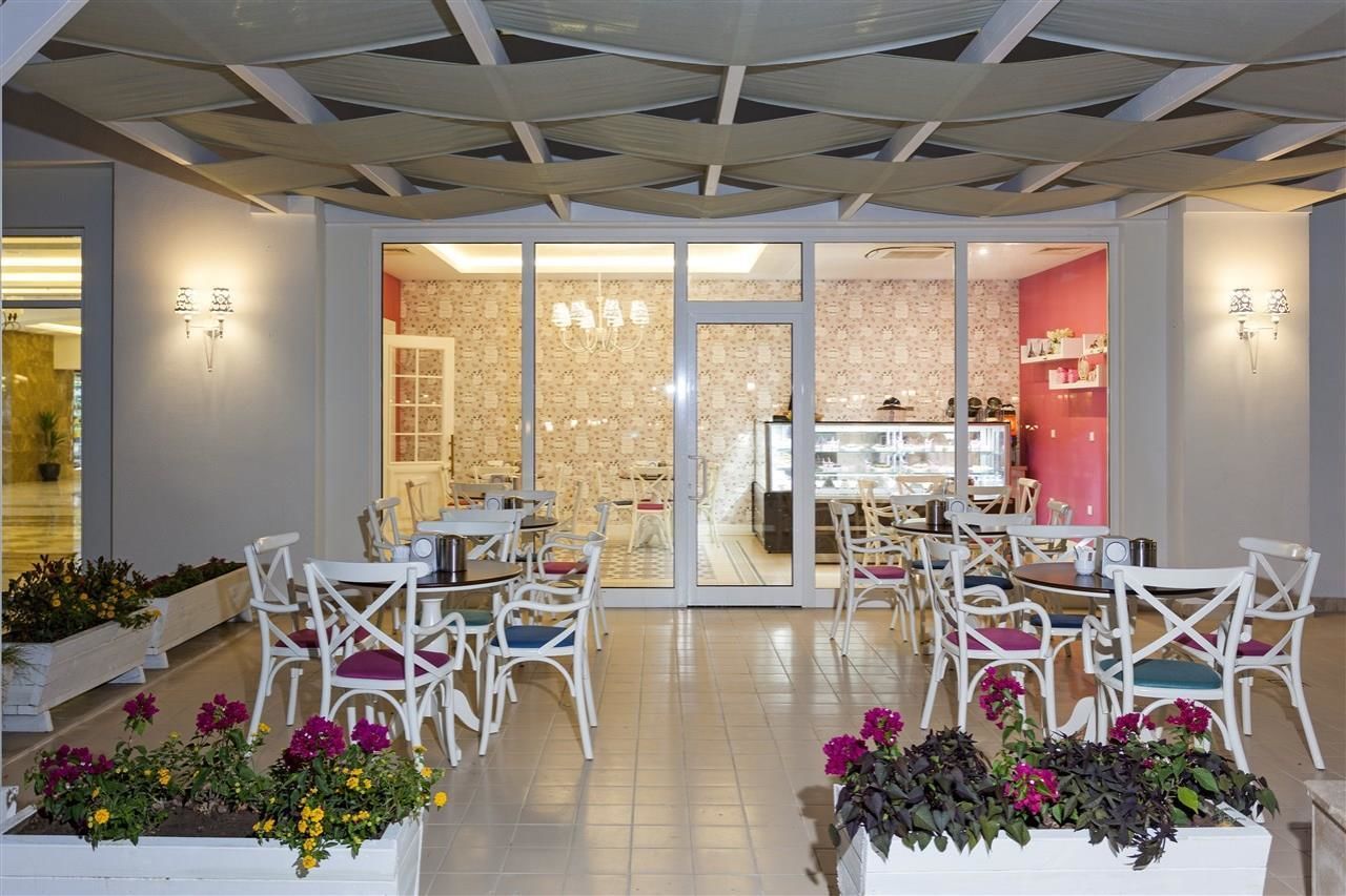 Un área de comedor interior moderna con sillas y mesas blancas, un divisor de vidrio y una cocina visible a través del mismo.
