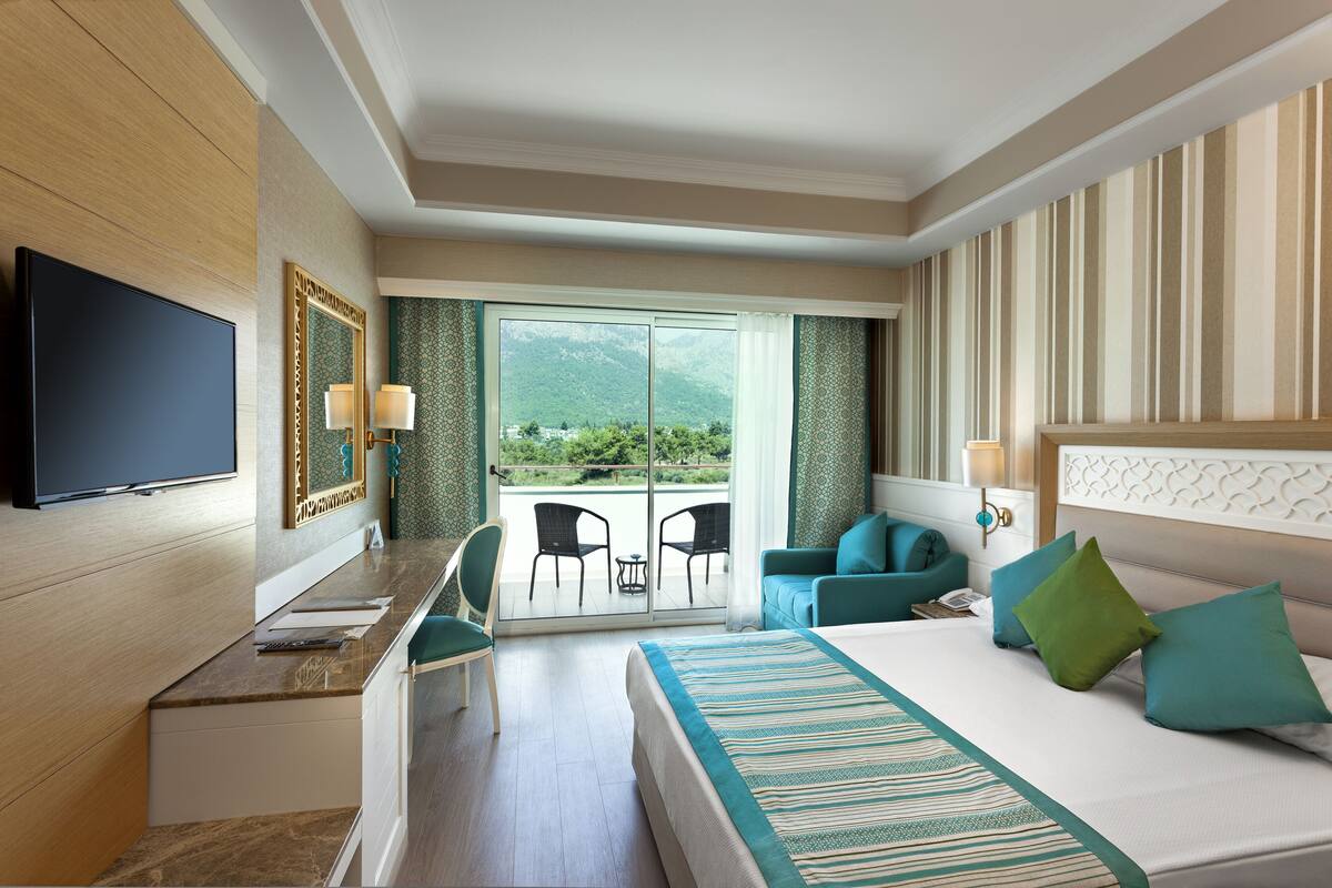 A hotel room with a bed, a desk, a TV, and a balcony with chairs.